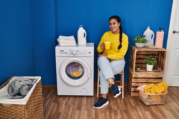 Young hispanic woman drinking coffee waiting for washing machine at laundry room