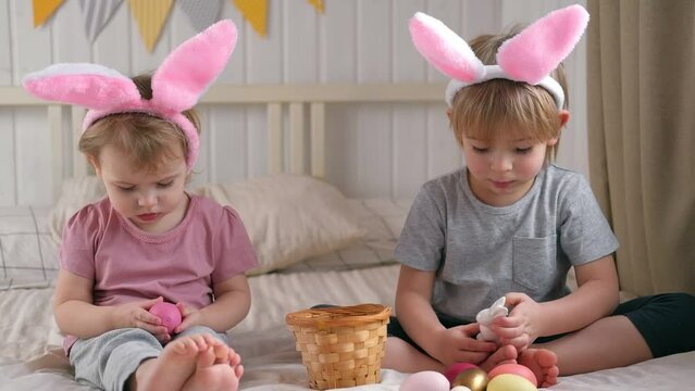 Happy children, baby girl and boy, wearing bunny ears headband playing at home in bedroom with colored Easter eggs. Siblings Enjoying the holidays. Easter day. Christian Passover. Celebrating Easter.
