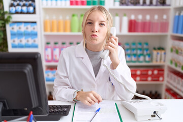 Young caucasian woman working at pharmacy drugstore speaking on the telephone looking at the camera blowing a kiss on air being lovely and sexy. love expression.