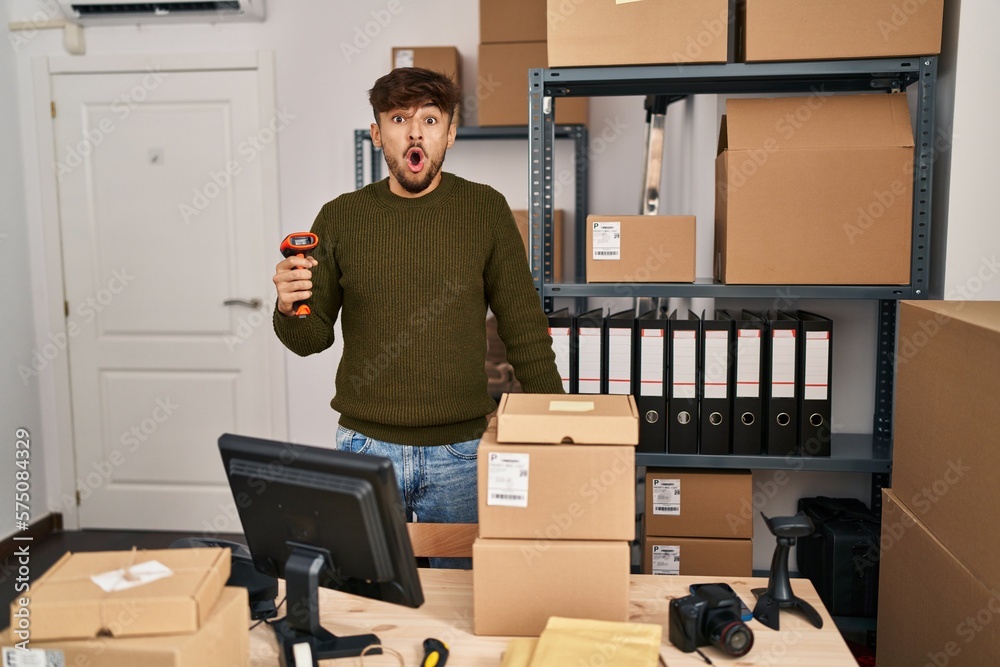 Canvas Prints Arab man with beard working at small business commerce holding bar code scanner scared and amazed with open mouth for surprise, disbelief face