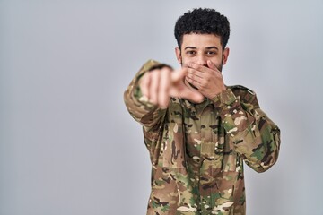 Arab man wearing camouflage army uniform laughing at you, pointing finger to the camera with hand over mouth, shame expression