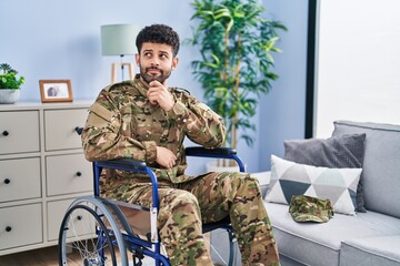 Fototapeta premium Arab man wearing camouflage army uniform sitting on wheelchair with hand on chin thinking about question, pensive expression. smiling with thoughtful face. doubt concept.