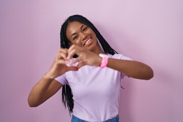 African american woman with braids standing over pink background smiling in love doing heart symbol shape with hands. romantic concept.