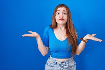 Redhead woman standing over blue background clueless and confused expression with arms and hands raised. doubt concept.