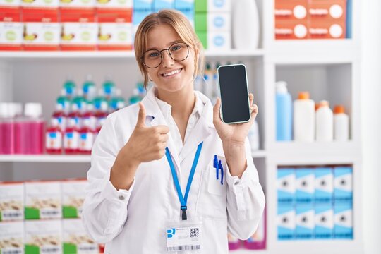 Young caucasian woman working at pharmacy drugstore showing smartphone screen smiling happy and positive, thumb up doing excellent and approval sign