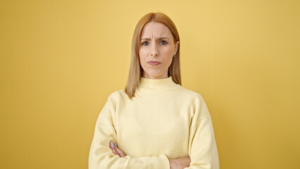 Young blonde woman standing with relaxed expression and arms crossed gesture over isolated yellow background