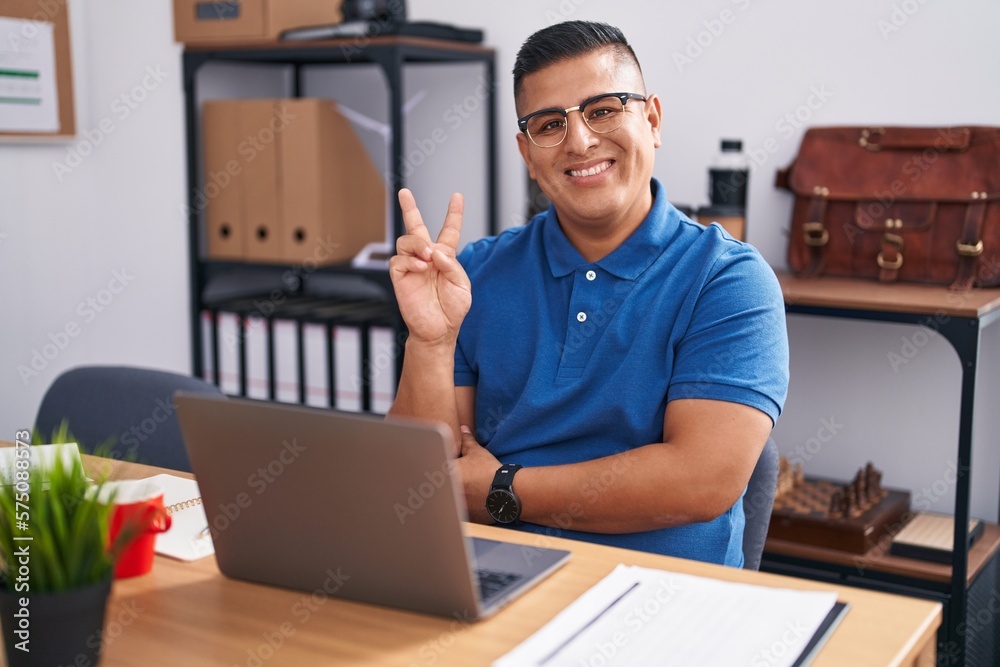 Sticker Young hispanic man working at the office with laptop smiling with happy face winking at the camera doing victory sign. number two.