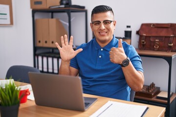 Young hispanic man working at the office with laptop showing and pointing up with fingers number six while smiling confident and happy.