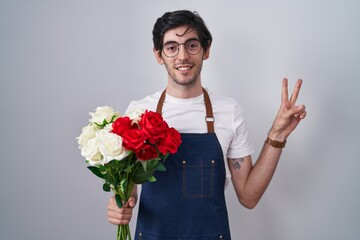 Young hispanic man holding bouquet of white and red roses smiling with happy face winking at the camera doing victory sign with fingers. number two.