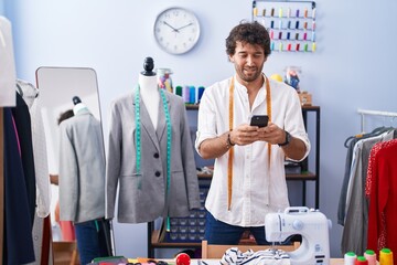 Young hispanic man tailor smiling confident using smartphone at clothing factory