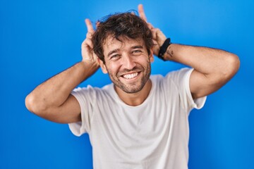 Fototapeta na wymiar Hispanic young man standing over blue background posing funny and crazy with fingers on head as bunny ears, smiling cheerful