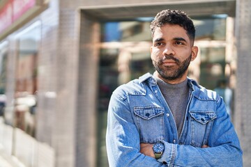 Naklejka premium Young hispanic man with relaxed expression standing with arms crossed gesture at street
