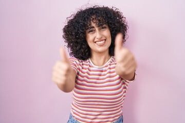 Young middle east woman standing over pink background approving doing positive gesture with hand, thumbs up smiling and happy for success. winner gesture.
