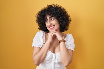 Fototapeta na wymiar Young brunette woman with curly hair standing over yellow background laughing nervous and excited with hands on chin looking to the side