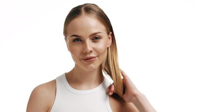 Good-looking fit European young woman with long blond hair in ponytail in white top turns to the camera touching her face and smiling wide for it on white background