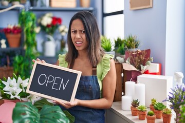 Hispanic young woman working at florist with open sign clueless and confused expression. doubt...