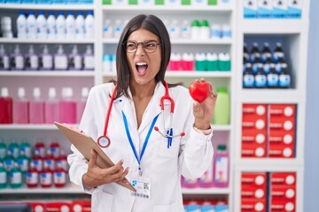 Brunette young woman working at pharmacy drugstore holding red heart angry and mad screaming frustrated and furious, shouting with anger. rage and aggressive concept.