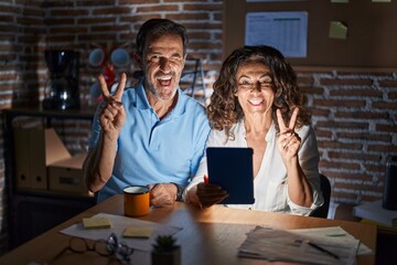 Middle age hispanic couple using touchpad sitting on the table at night smiling with happy face winking at the camera doing victory sign with fingers. number two.