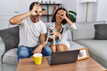 Hispanic middle age couple sitting on the sofa using computer laptop smiling happy doing ok sign with hand on eye looking through fingers