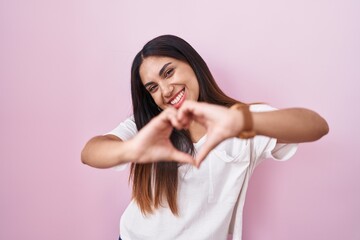 Young arab woman standing over pink background smiling in love doing heart symbol shape with hands. romantic concept.