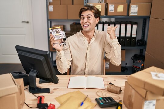 Young Man Working At Small Business Ecommerce Holding Cart Pointing Thumb Up To The Side Smiling Happy With Open Mouth