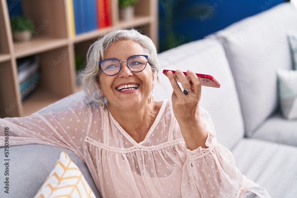 Poster Middle age grey-haired woman listening voice message by smartphone sitting on sofa at home