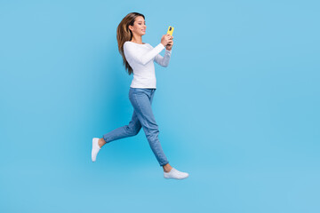 Fototapeta premium Full length photo of pretty shiny woman wear white shirt jumping high chatting device empty space isolated blue color background