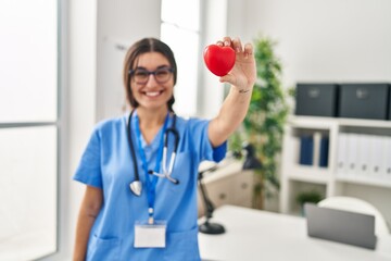 Young hispanic woman wearing doctor uniform holding heart at clinic