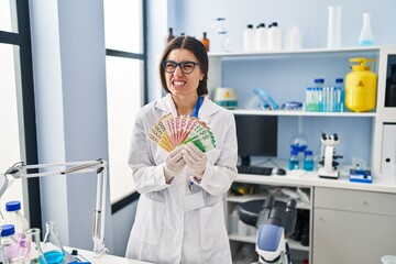 Young hispanic woman working at scientist laboratory holding money banknotes angry and mad...