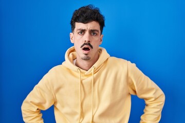 Hispanic man standing over blue background in shock face, looking skeptical and sarcastic, surprised with open mouth