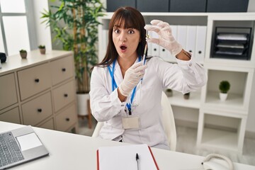 Young brunette doctor woman holding syringe and vaccine afraid and shocked with surprise and amazed expression, fear and excited face.