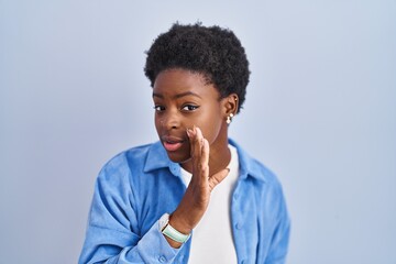 African american woman standing over blue background hand on mouth telling secret rumor, whispering malicious talk conversation