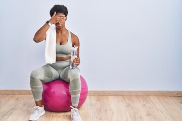 African american woman wearing sportswear sitting on pilates ball covering eyes with hand, looking...