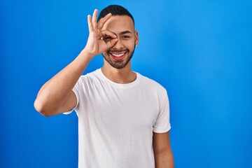 Naklejka premium Young hispanic man standing over blue background doing ok gesture with hand smiling, eye looking through fingers with happy face.