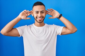 Young hispanic man standing over blue background smiling pulling ears with fingers, funny gesture....