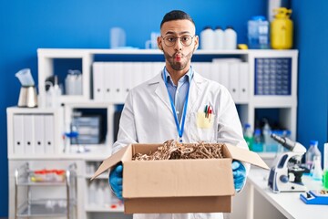 Young hispanic man working at scientist laboratory holding cardboard box making fish face with...