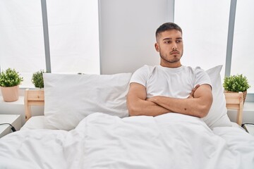 Young hispanic man with serious expression sitting on bed at bedroom