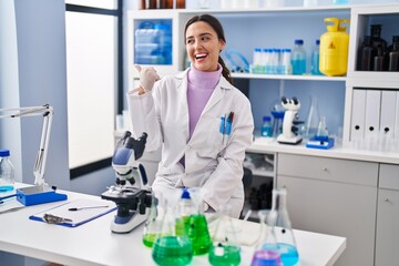 Fototapeta premium Young brunette woman working at scientist laboratory smiling with happy face looking and pointing to the side with thumb up.