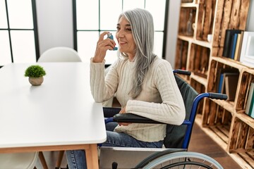 Middle age grey-haired woman using inhaler sitting on wheelchair at home