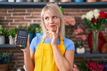 Young caucasian woman working at florist shop holding dataphone serious face thinking about...