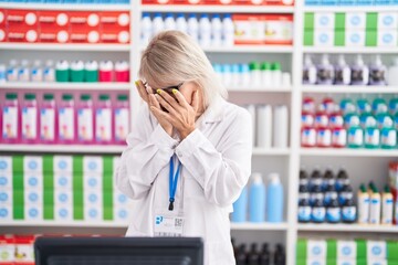 Young caucasian woman working at pharmacy drugstore with sad expression covering face with hands...