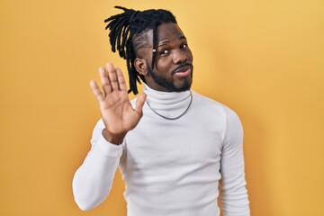 African man with dreadlocks wearing turtleneck sweater over yellow background waiving saying hello...
