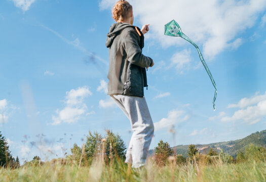 Teenager boy with flying green color kite on the high grass meadow in the mountain fields. Happy childhood moments or outdoor time spending concept image.