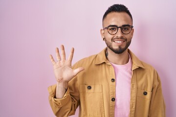 Young hispanic man standing over pink background showing and pointing up with fingers number five while smiling confident and happy.