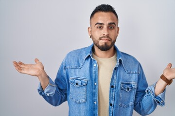 Young hispanic man standing over isolated background clueless and confused expression with arms and hands raised. doubt concept.