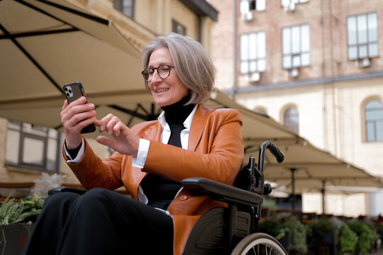 elder woman in wheelchair holding mobile phoneoutdoor. elderly female texting message, using app with cellphone in park. - Powered by Adobe