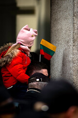 People in Lithuania celebrating their independence day gathered together with Lithuanian flags; a small girl sitting on man shoulders with a flag