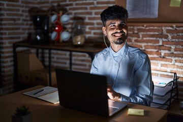 Young hispanic man with beard working at the office at night looking away to side with smile on face, natural expression. laughing confident.