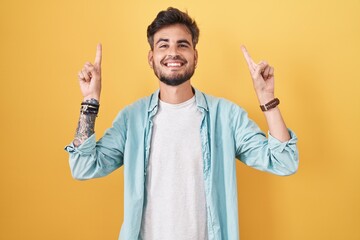 Young hispanic man with tattoos standing over yellow background smiling amazed and surprised and pointing up with fingers and raised arms.