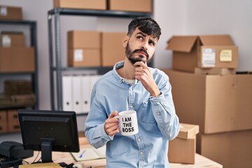 Young hispanic man with beard working at small business ecommerce drinking from boss cup serious...